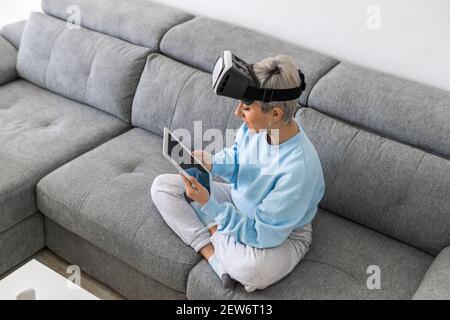 An adult woman using a VR headset and a tablet while sitting on a sofa Stock Photo