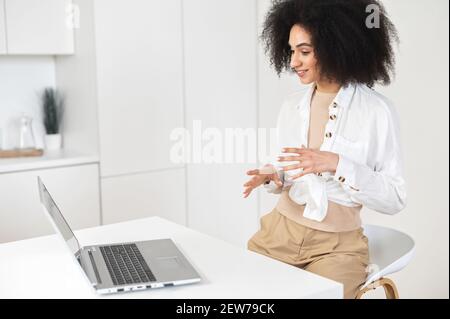 Stylish African American female entrepreneur businesswoman with Afro hairstyle sitting in the kitchen and communicating online via laptop, gesturing and discussing project or checking up on a friend Stock Photo