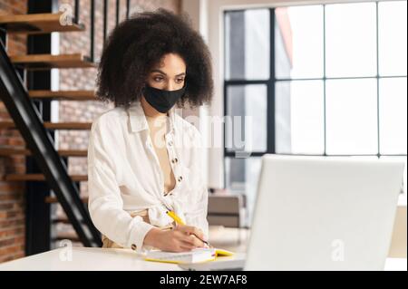 African American female student or businesswoman with curly hair wearing black protective medical face mask sitting at desk in coffee shop or library, working on laptop, taking notes to notebook Stock Photo