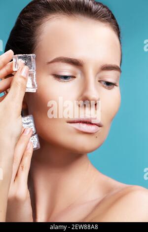 Closeup portrait of beautiful young woman applies the ice to face on a blue background. Skin care concept. Stock Photo