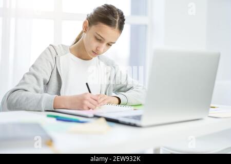 Concentrated latin teenage girl making notes in her notepad, using laptop while having online lesson at home. Distance education, home schooling concept Stock Photo