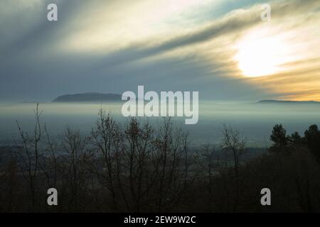 beautiful landscape showing the sun over a layer of fog with some trees and a mountain in the background Stock Photo