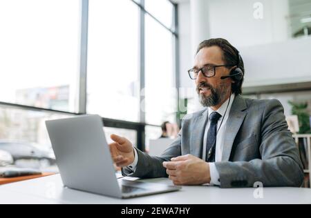 Senior adult business male in headset is sitting at the desk, using laptop, communicate with clients. Caucasian grey-haired manager is consulting people, support service concept Stock Photo