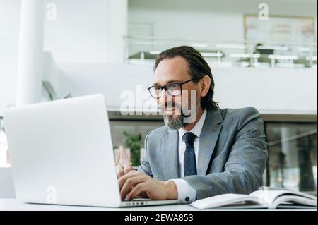 Smiling mature male employee is chatting with colleagues uses a laptop. Successful businessman working on new project, searching ideas, browsing internet Stock Photo