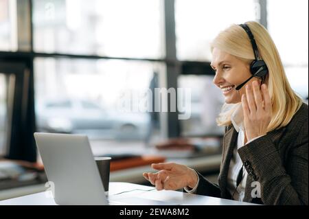 Caucasian female employee in headset is using laptop, sitting at the desk. Senior happy business woman talking with video colleagues at online conference, smiling, discussing ideas Stock Photo
