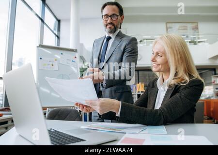 Successful senior coworkers developing new project, discussing ideas. Beautiful blonde-hair woman working on business plan together with attractive man colleague Stock Photo
