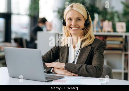 Middle aged woman manager in headset and stylish elegant suit is looking at the camera, smiling. Happy successful female operator of call center is sitting at the desk in office Stock Photo