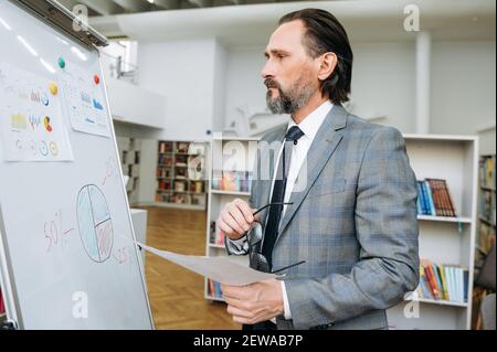 Serious influential businessman is learning financial graphs, standing near flipchart in office. Concentrated senior male developing important project, looks at the marker board Stock Photo