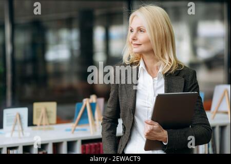 Stylish elegant mature lady in formal wear standing in office, looking at the side, thinking about new project. Gorgeous business woman is working as office employee, holding laptop in arms Stock Photo