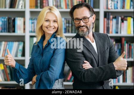 Friendly senior adult colleagues posing at the workplace, showing thumbs up. Elegant pretty woman with handsome gray-haired man looking at the camera and smiling, partnerships concept Stock Photo