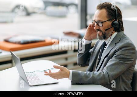 Handsome senior adult businessman in headset is using laptop, having a video conference. Happy male manager talking with coworkers, smiling, discussing corporate project Stock Photo