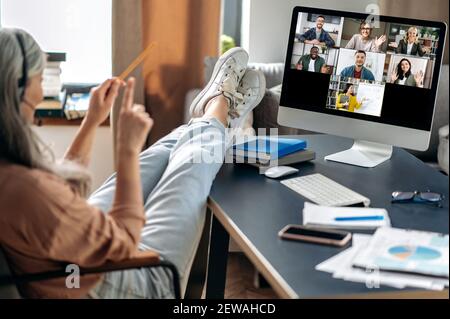 Senior gray-haired woman sits at home at a table with her legs thrown on the table, listens to an online lesson by video conference using computer, teacher and students are on the screen, distance Stock Photo