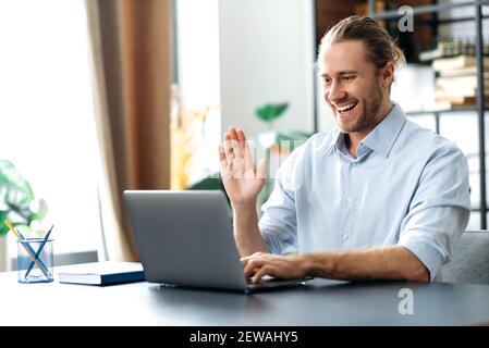 Attractive freelancer guy using laptop, have a video call, communicate with colleagues or friends by video conference, waving hand and smiling. Handsome caucasian man sits at the desk, working from Stock Photo