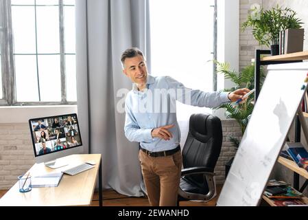 Middle aged successful male top manager or business tutor standing near flipchart in modern office, conducts an online lesson or presentation. Satisfied businessman stylishly dressed on video meeting Stock Photo
