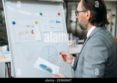 Middle aged businessman looking at the flipchart, learning work reports. Focused male employee is working on new business plan for a company Stock Photo