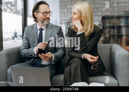 Friendly mature business partners take a break from work, talking about different news. Happy colleagues sitting on the sofa in stylish suits, having a conversation, communication concept Stock Photo