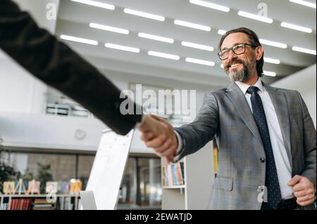 Successful grey-haired male leader of the company is shaking hands with business partner on meeting. Influential colleagues coming to agreement on negotiations, sign corporate contract Stock Photo