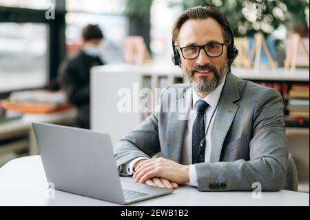 Mature successful grey-haired man in headset and stylish suit looks at the camera, smiling. Happy successful male operator of call center sits at the desk in office, consulting people Stock Photo