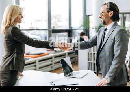 Successful middle aged colleagues are standing in modern office, coming to agreement at conference. Satisfied business partners shaking hands after negotiations Stock Photo