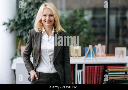 Confident middle aged business woman in stylish suit posing at the workplace. Successful happy elegant lady looking directly at the camera, smiling, working as office employee Stock Photo