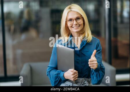 Portrait of happy caucasian charming business lady in eyeglasses at the workplace, showing thumbs up, holds a laptop, looking at the camera with friendly smile Stock Photo