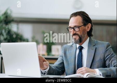 Joyful middle aged businessman is using laptop, having online conference. Successful male employee talking by video call with colleagues, discussing ideas Stock Photo