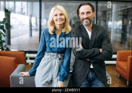 Happy caucasian middle aged business people in stylish wear are standing in modern office, looking at the camera, smiling, partnership concept Stock Photo
