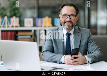 Portrait of a confident senior business man in formal suit and eyeglasses is sitting at the workplace, looking and friendly smiling directly at the camera Stock Photo