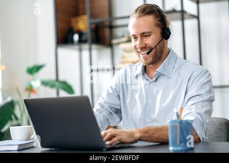 Joyful young adult male manager or call center worker in headset is looks at the laptop screen, having online business meeting. Successful employee talking online with colleagues Stock Photo