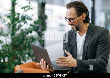 Joyful stylish businessman on video conference with coworkers, discussing corporate project. Handsome male employee using laptop, sitting on sofa in modern office, smiling Stock Photo