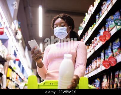 Young black woman in face mask holding smartphone, going through shopping list at groceries department of supermarket Stock Photo