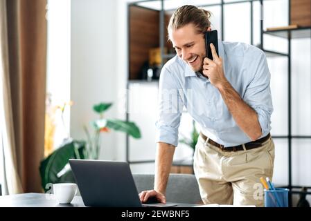 A very busy caucasian successful businessman or freelancer working from home, talking on the phone with a colleague while using a laptop, browsing the internet, answering an email Stock Photo