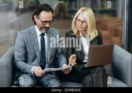 Concentrated caucasian senior colleagues working together on new business strategy for company. Serious male and female employees learning documents, using laptop, sitting on the sofa Stock Photo