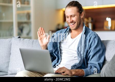 Joyful male freelancer using laptop, have a video call, communicate with colleagues or friends by video conference, waving hand and smiling. Handsome caucasian man is sitting on a sofa, working from Stock Photo
