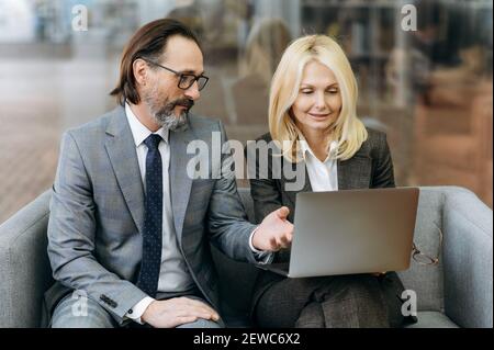 Middle aged focused coworkers are sitting on the sofa, using laptop. Caucasian colleagues in formal suits working on business plan, searching ideas, browsing internet, partnership concept Stock Photo