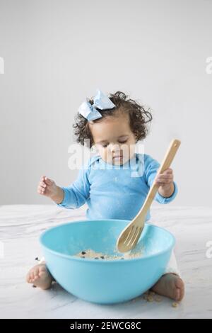 A little girl sitting on a counter stirring cookie dough mix with a wooden spoon. Stock Photo