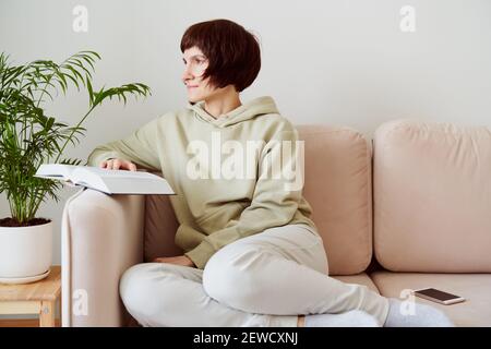 Mature woman reading book sitting on sofa, looking to window, dreaming and thinking. Digital detox Stock Photo
