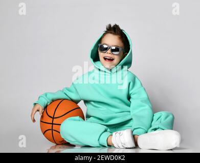 Laughing loudly frolic kid boy in modern green, mint color sportswear hoodie and pants sits on floor with basketball Stock Photo