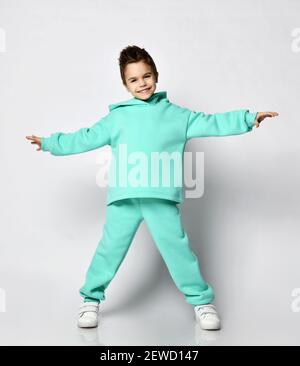 Young active positive kid boy with stylish haircut in modern green, mint color hoodie stands holding hands up spread Stock Photo