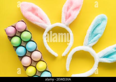 Eggs hunting concept. Easter holiday flat lay. Colored Easter eggs in egg carton nd bunny ears on yellow background. Stock Photo