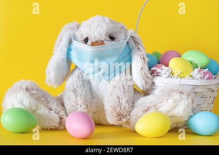 Easter stuffed toy bunny in protective face mask and colored eggs on yellow background. Ester holiday during coronavirus pandemic concept. Stock Photo