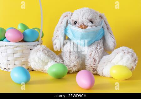 Easter stuffed toy bunny in protective face mask and colored eggs on yellow background. Ester holiday during coronavirus pandemic concept. Stock Photo