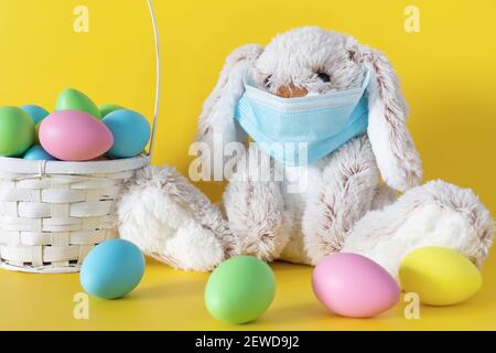 Easter stuffed toy bunny in protective face mask and colored eggs on yellow background. Ester holiday during coronavirus pandemic concept. Stock Photo