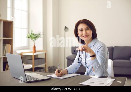 Happy businesswoman sitting at desk with laptop, taking notes and smiling at camera Stock Photo