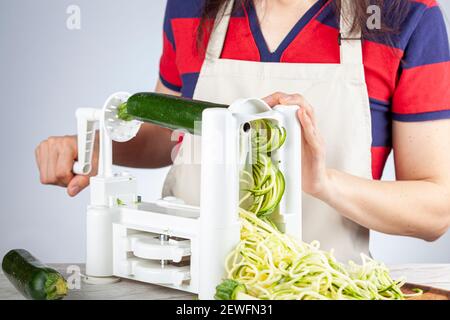 A caucasian woman wearing apron is turning the handle of a vegetable spiralizer, slicer to make homemade zucchini noodles on kitchen countertop. This Stock Photo