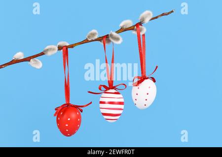 Red and white Easter eggs with stripes and dots hanging from goat willow branch on blue background Stock Photo