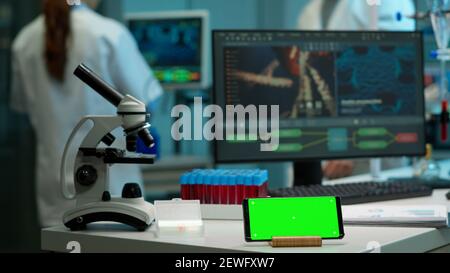 Microbiologist bringing blood samples in modern lab putting them near smartphone working with green chroma key screen placed on desk. Team of biotechnology scientists developing drugs in background Stock Photo