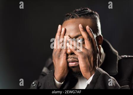 Scared black man covering his face with his hands, looking at the camera with the only eye that is not covered by his fingers. Close up. Black backgro Stock Photo