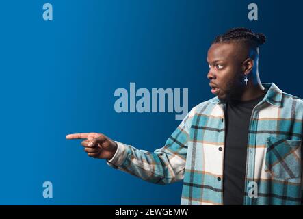 Handsome black man pointing aside, surprised. Mid shot. Blue background. Stock Photo