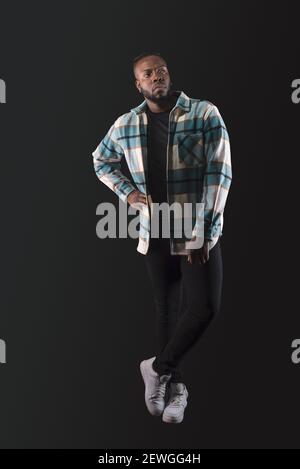 Handsome black man, standing looking serious to the side, wearing blue plaid shirt and jeans, legs crossed. Full body. Blue background. Stock Photo